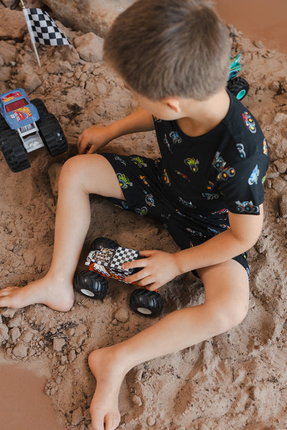 Boy sitting on sand and rocks with toy trucks wearing Black Toddler Basic Tee and drawstring short in Monster Truck print