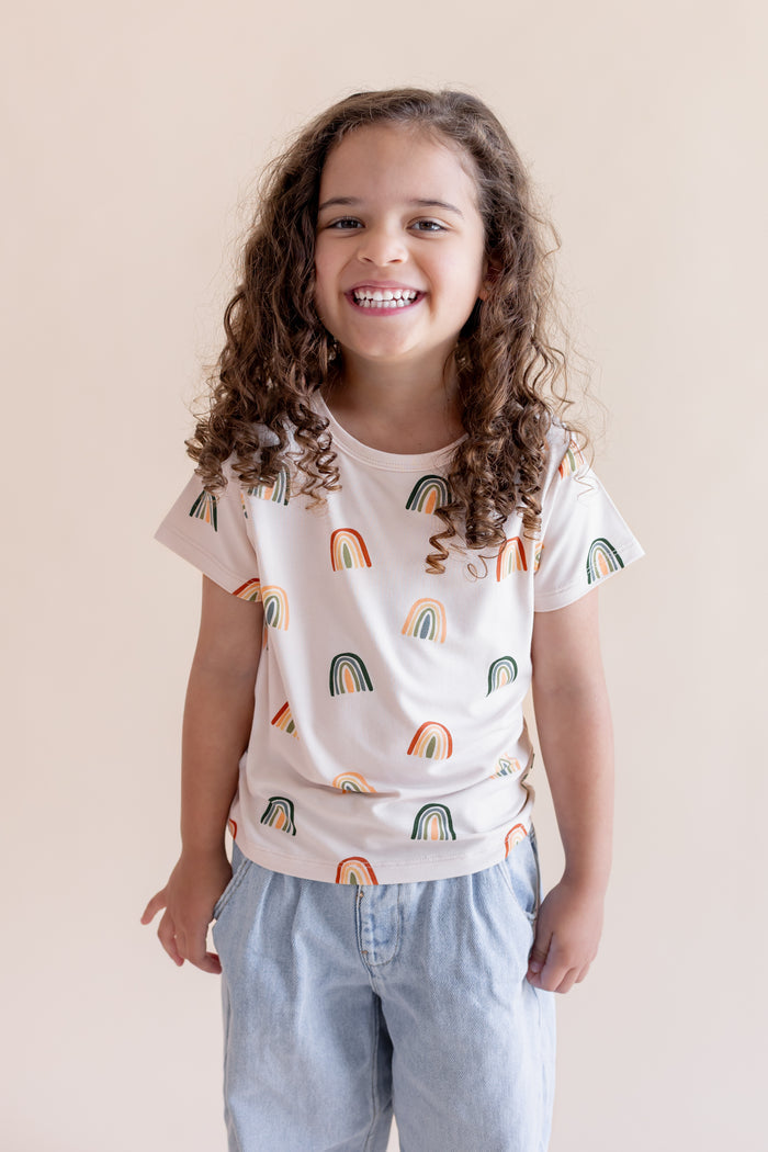 Girl posing in Toddler Basic Tee in Retro Rainbow print over blue pants on a white background
