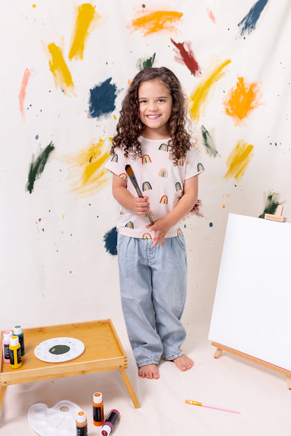 Girl posing in Toddler Basic Tee in Retro Rainbow print over blue pants holding a paint brush in front of a colorful background with few paints and canvas around her