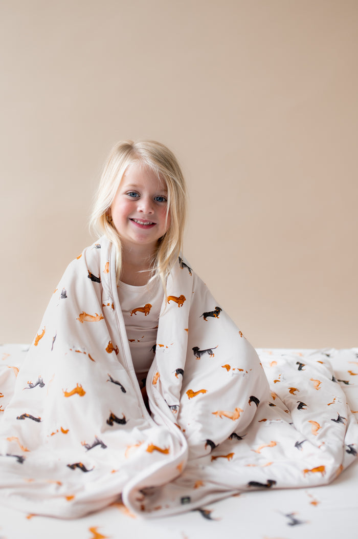 Girl wrapped in a Beige Toddler Blanket in Dachshund print sitting on a similar print sheet.