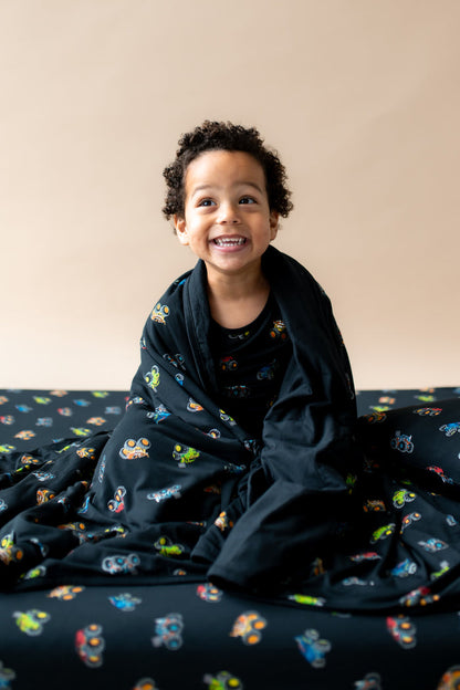 Boy wrapped in black Toddler Blanket in Monster Truck print sitting on similar print sheet.