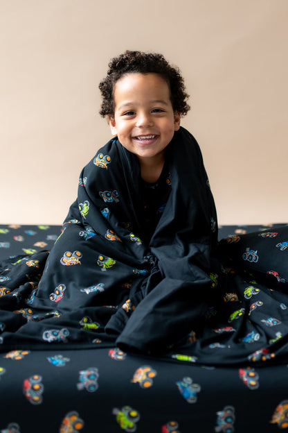 Boy smiling wrapped in black Toddler Blanket in Monster Truck print sitting on similar print sheet.