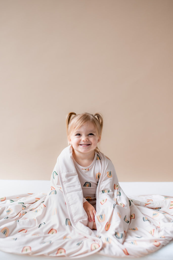 Girl sitting wrapped in Toddler Blanket in Retro Rainbow 1.0 print wearing a similar print long sleeve pajama set on a beige background