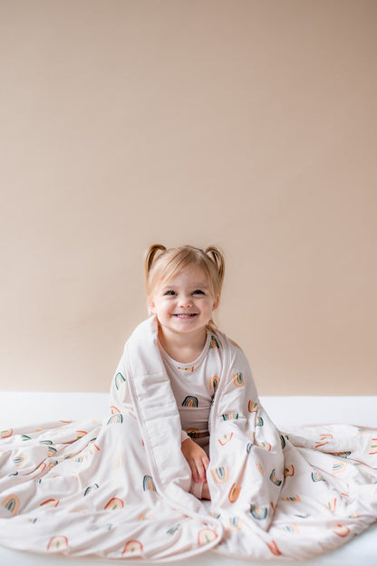 Girl sitting wrapped in Toddler Blanket in Retro Rainbow 1.0 print wearing a similar print long sleeve pajama set on a beige background
