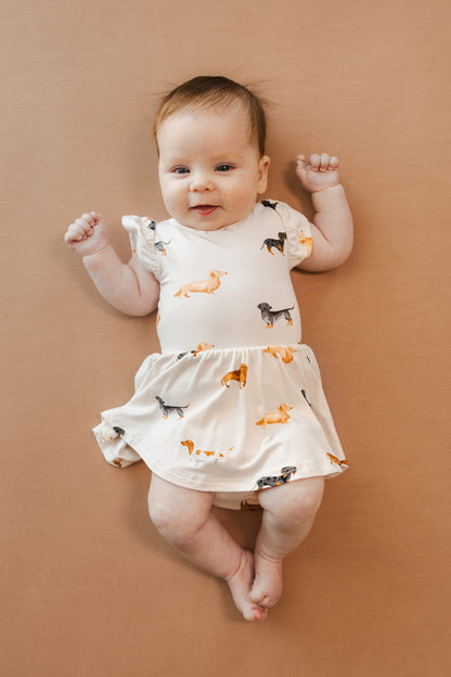 Baby wearing Beige Twirl Bodysuit Dress in Dachshund print on a brown background
