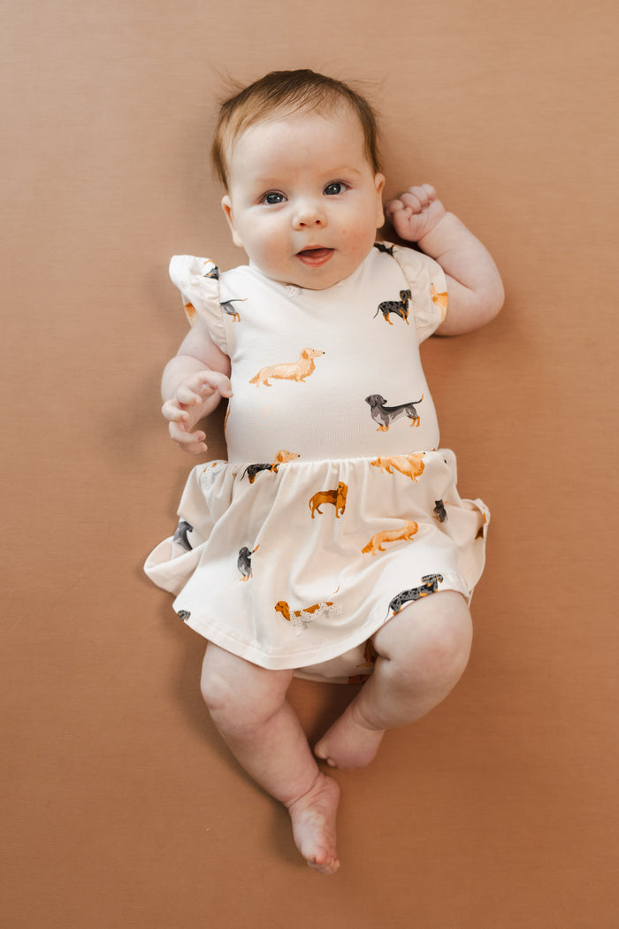 Baby in Beige Twirl Bodysuit Dress in Dachshund print on a brown background