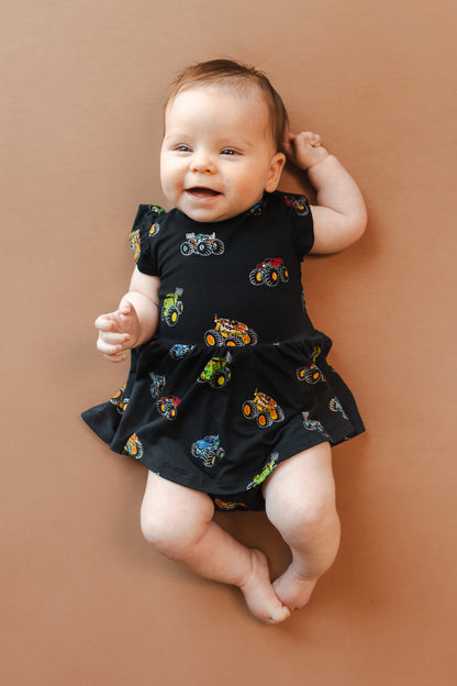 Baby wearing Black Twirl Bodysuit Dress in Monster Truck print on beige background