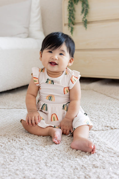 Baby sitting on a white carpet wearing Twirl Bodysuit Dress in Retro Rainbow print