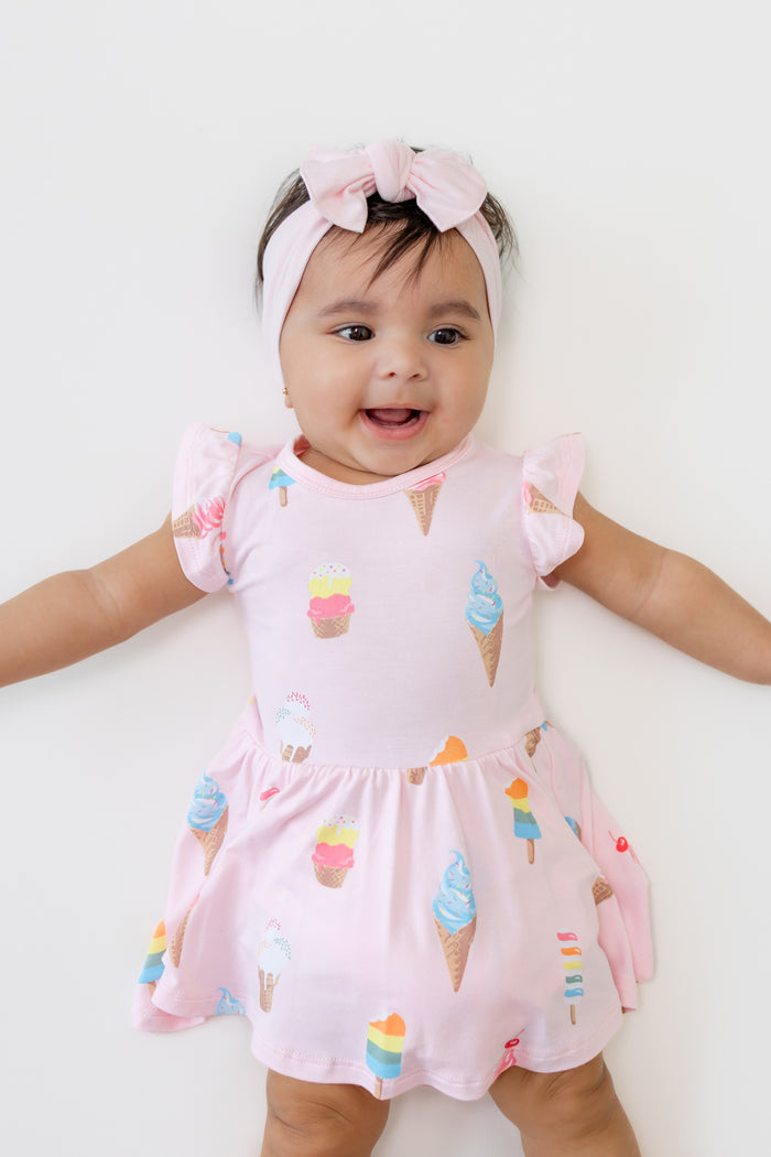 Baby wearing Twirl Bodysuit Dress in Sakura Ice Cream print with a pink bow headband on a beige background