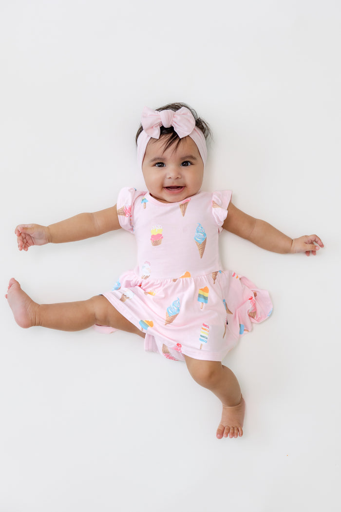 Baby posing in Twirl Bodysuit Dress in Sakura Ice Cream print wearing a pink bow headband on a beige background