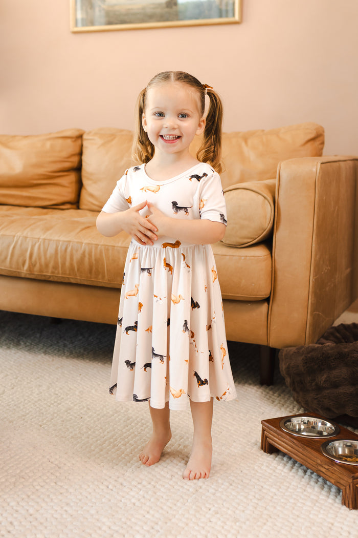 Girl wearing Beige Twirl Dress in Dachshund print standing in front of beige couch in a living room