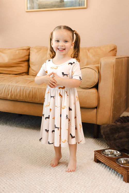 Girl wearing Beige Twirl Dress in Dachshund print standing in front of beige couch in a living room