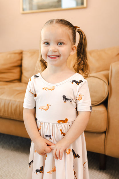 Girl smiling in Beige Twirl Dress in Dachshund print standing in front of beige couch in a living room