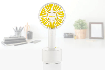 Ein kompaktes weiß-gelbes Unold Ventilatoren-Set Tischventilator Breezy Clip + Handventilator Breezy Swing steht auf einem Schreibtisch, mit verschwommenen Büromaterialien, einer kleinen Pflanze und einem Bilderrahmen im Hintergrund.