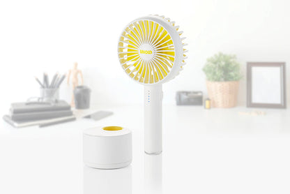 Das Unold Ventilatoren-Set Tischventilator Breezy Clip + Handventilator Breezy Swing in Weiß und Gelb steht aufrecht auf einer weißen Fläche neben seiner runden Ladestation, im Hintergrund sind unscharfe Büromaterialien und eine Pflanze zu sehen.