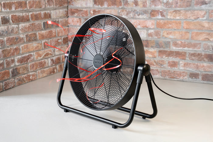 Der elektrische Bodenventilator Unold Windmaschine Black Steel, der nach oben gerichtet ist, bläst rote Bänder, um den Luftstrom in einem Raum mit hellgrauem Fußboden und Sichtziegelwänden zu zeigen. Der schwarze Ventilator ist eingesteckt.