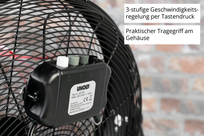 Nahaufnahme des schwarzen Schaltkastens der Unold Windmaschine Black Steel auf einem Gebläsegitter, mit drei beschrifteten Knöpfen für die 3-Stufen-Regelung und einem Tragegriff. Ein deutscher Text erklärt die Funktionen; im Hintergrund ist eine unscharfe Backsteinmauer zu sehen.