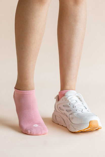 A close-up of a person's lower legs and feet, showing one ankle in a light pink no-show sock and the other foot wearing a white athletic sneaker with a pink collar and orange outsole, against a solid pale beige background.