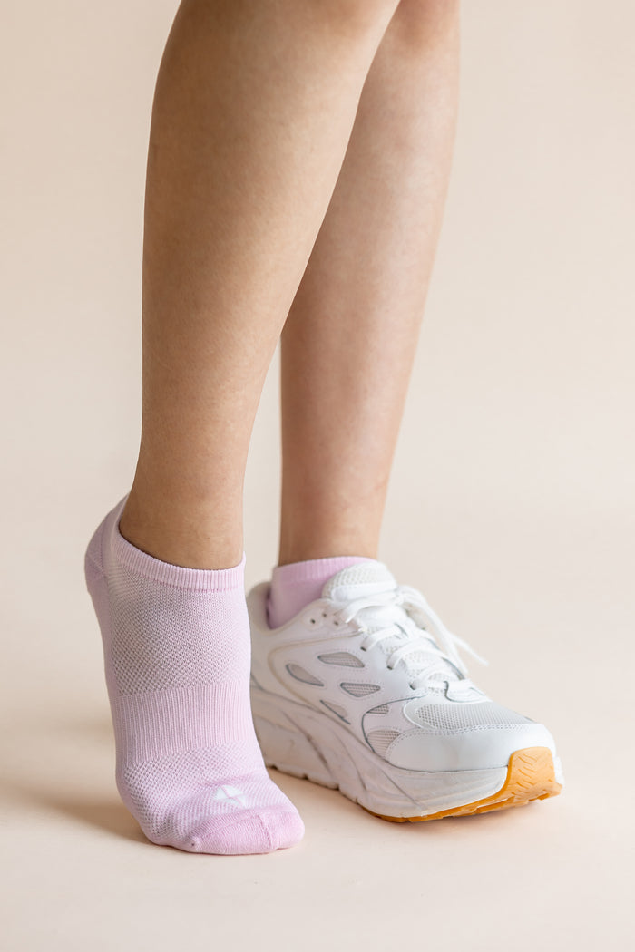 A close-up of a person's legs wearing a pair of light pink low-cut athletic socks and white sneakers with orange soles, set against a solid beige background.