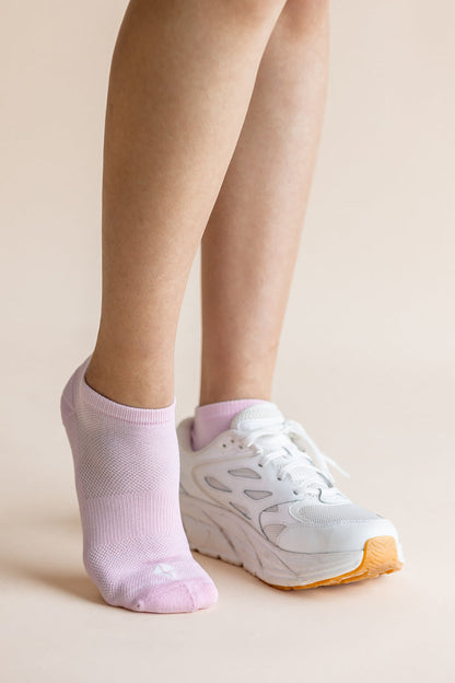 A close-up of a person's legs wearing a pair of light pink low-cut athletic socks and white sneakers with orange soles, set against a solid beige background.