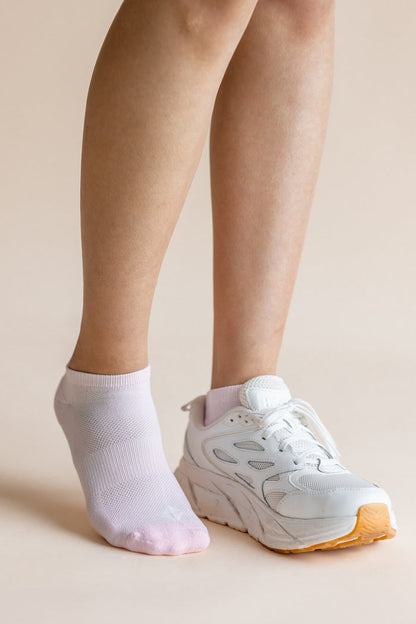 A person's legs and feet shown wearing a pair of white ankle socks, with one foot in a white sneaker and the other barefoot, against a solid beige background.