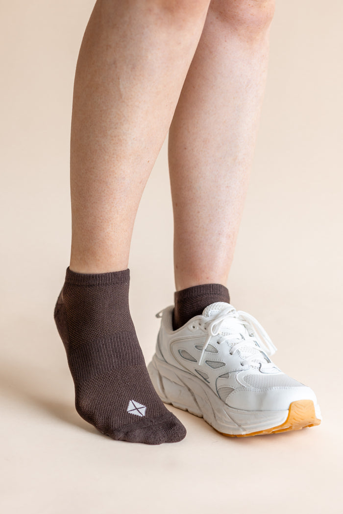A person's legs and feet shown wearing a dark brown ankle sock on one foot and a white athletic sneaker on the other, against a plain beige studio background.