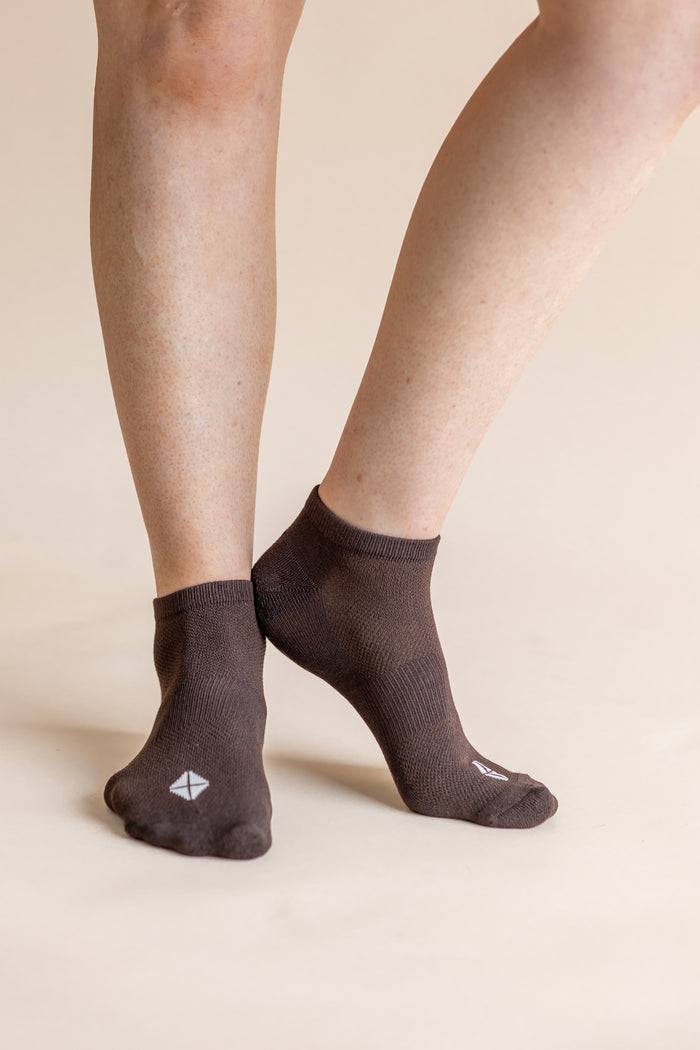 A close-up of a person's feet wearing dark brown low-cut socks with a small white geometric logo on the toes, set against a solid beige background.