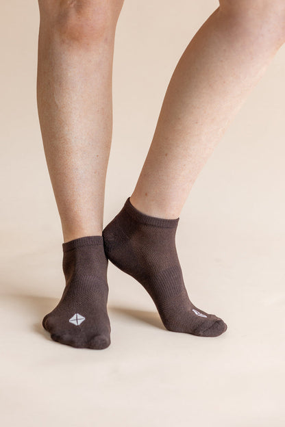 A close-up of a person's feet wearing dark brown low-cut socks with a small white geometric logo on the toes, set against a solid beige background.