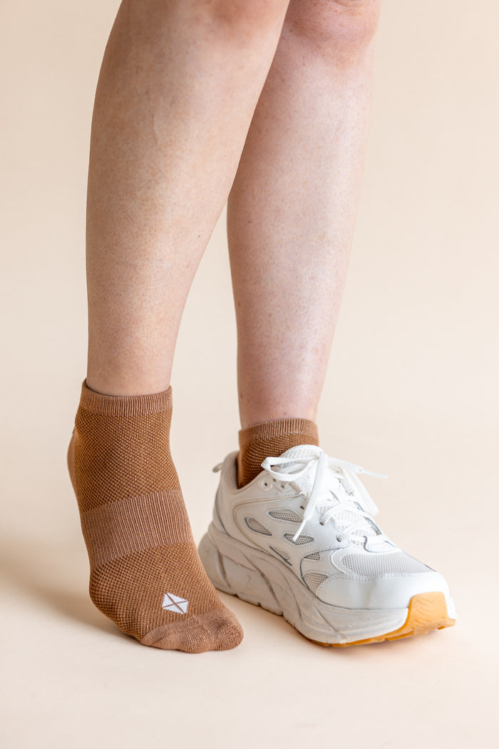 A close-up of a person's legs wearing a pair of low-cut brown knit socks and a white athletic sneaker against a solid beige background.