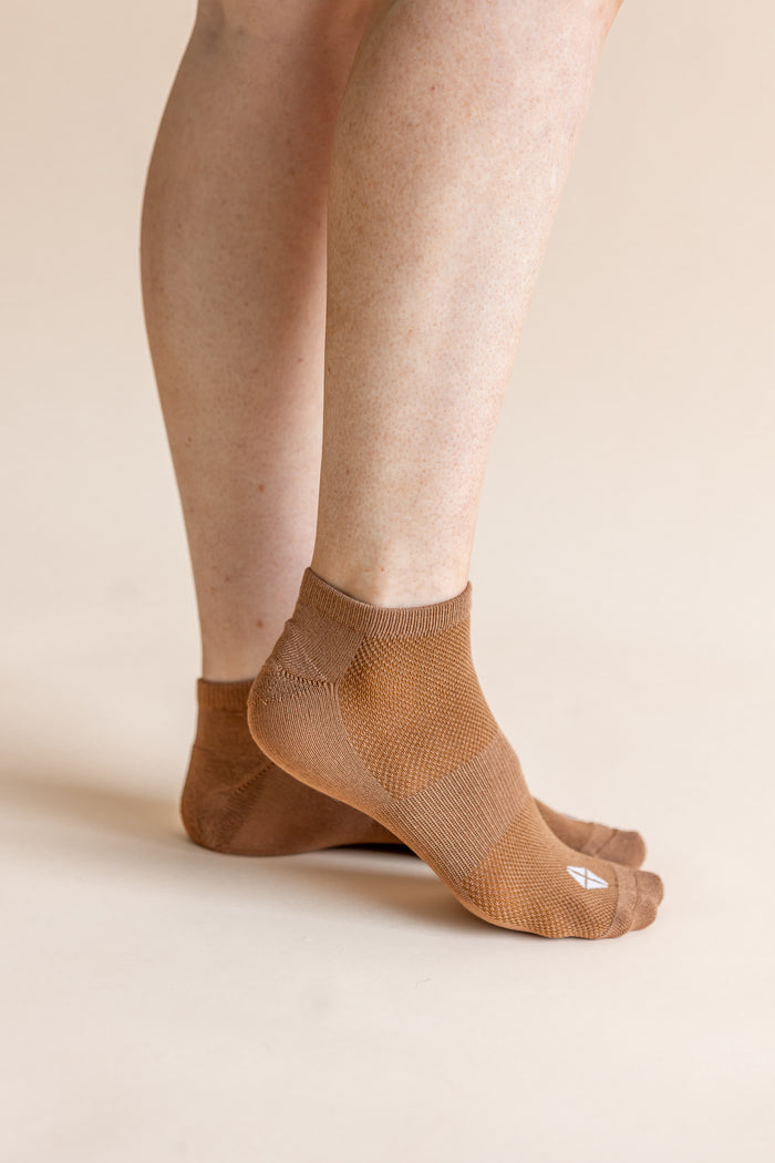 A close-up of a person's feet wearing tan low-cut athletic socks against a solid light beige background.