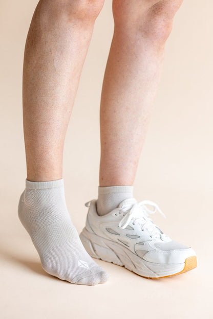 A close-up of a person's lower legs and feet, showing one ankle in a light gray ribbed ankle sock and the other in a white athletic sneaker, against a solid pale peach background.