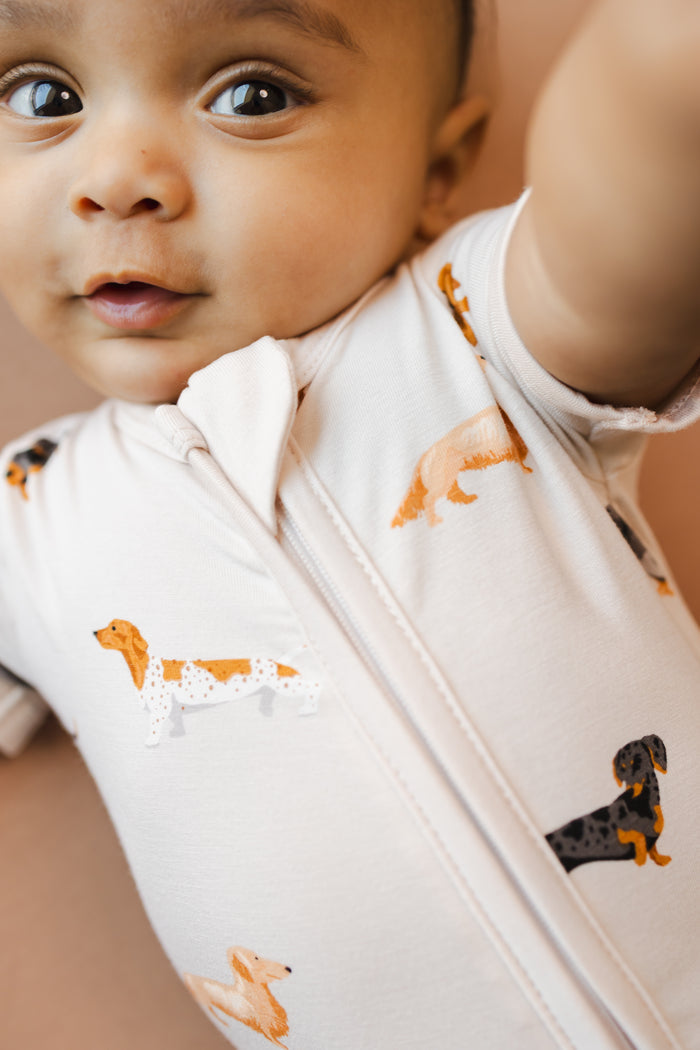 Close up of baby wearing Beige Zipper Shortall Romper in Dachshund print on a brown background