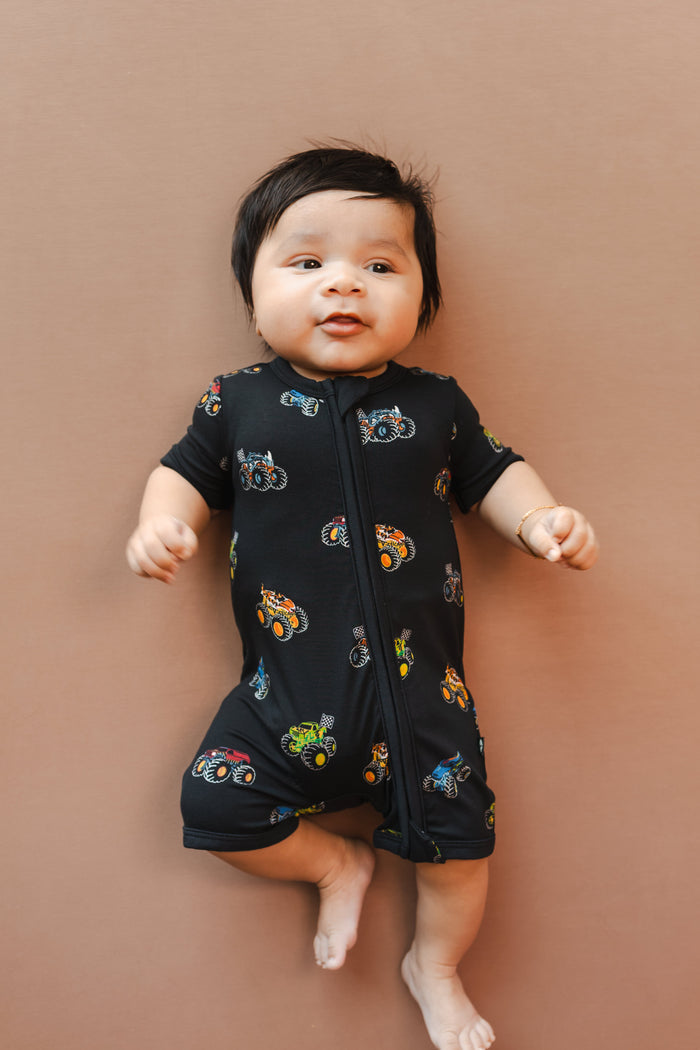 Baby wearing Black Zipper Shortall Romper in Monster Truck print on beige background