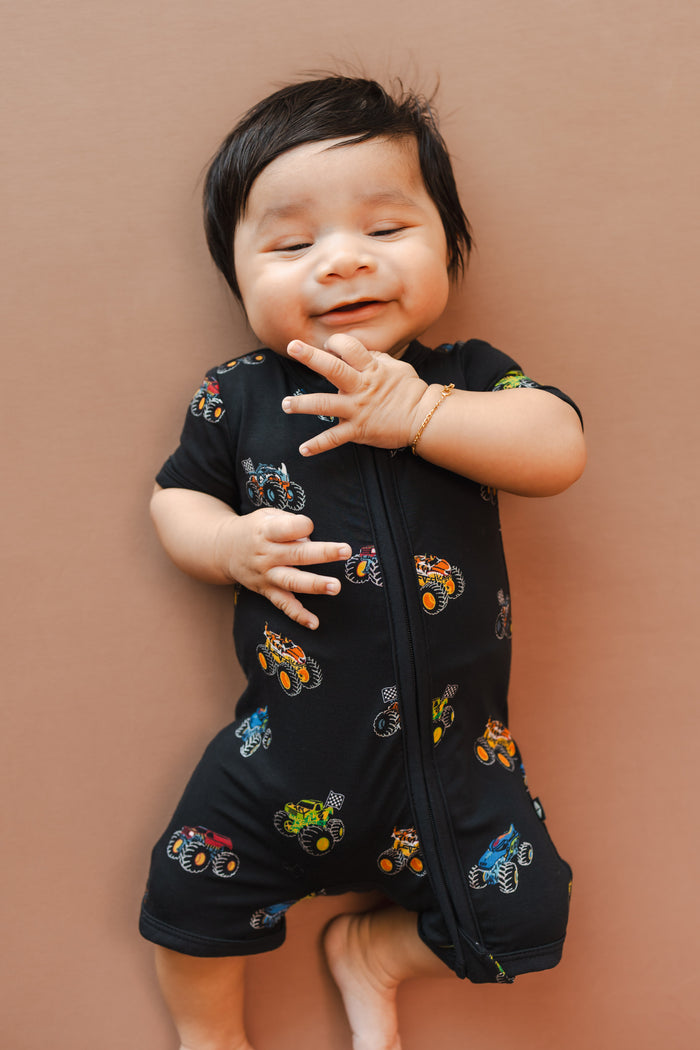 Baby in Black Zipper Shortall Romper in Monster Truck print on beige background