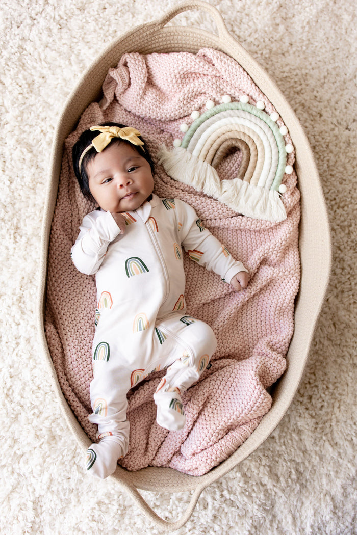 Baby lying in a basket over a pink blanket wearing Zippered Footie in Retro Rainbow print with a retro rainbow prop