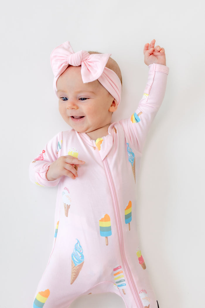 Baby lying in Sakura Ice cream print zippered footie wearing pink a bow headband on a white background