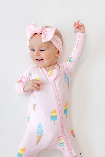 Baby lying in Sakura Ice cream print zippered footie wearing pink a bow headband on a white background