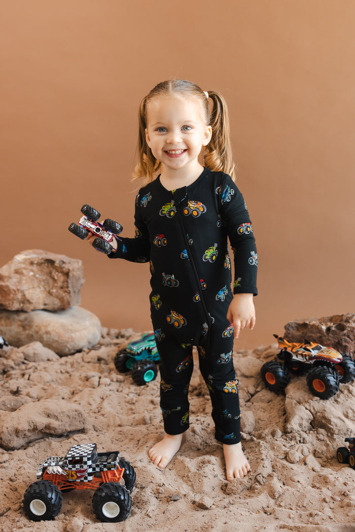 Girl holding toy truck wearing black Zippered Romper in Monster Truck print on sand and rocks with toy trucks