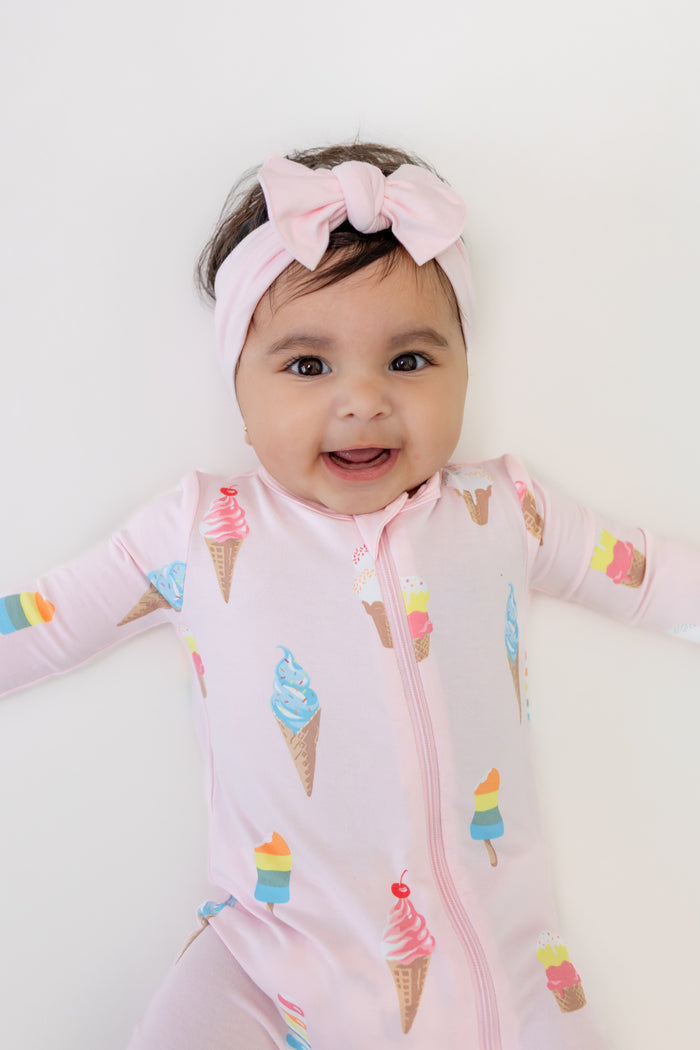 Baby lying in Zippered romper in Sakura Ice cream print with pink bow headband on a white background